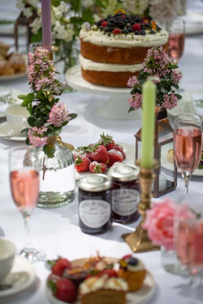 a beautifully arranged outdoor table near Solihull, featuring a layered sponge cake with berries, fresh strawberries, jam jars, pink drinks, and floral decorations.