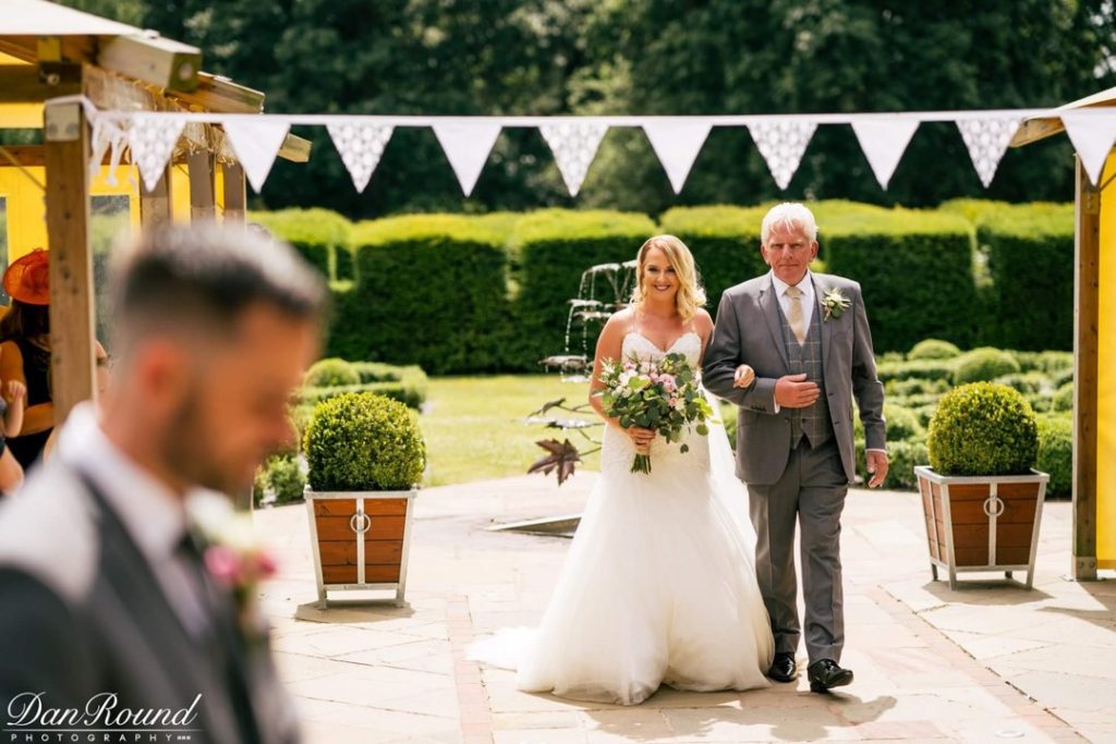 Bride walking down the aisle with her father under a bunting arch at an outdoor Stone Manor wedding.